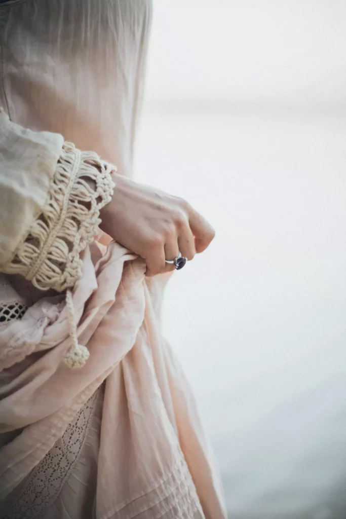 Close-up of a hand wearing a ring with a dark stone, holding gathered fabric. The fabric is light-colored and has lace detailing. The background is a blurred, light surface.