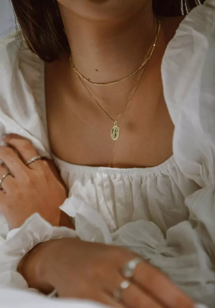 Close-up of a woman's chest wearing a white blouse and two gold necklaces. The shorter necklace is a delicate gold chain, and the longer necklace features a small, oval-shaped pendant with intricate detailing.
