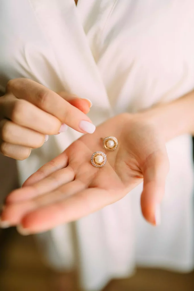 A woman in a white outfit holds a pair of gold pearl earrings with diamond accents in her palm, showcasing their intricate design with soft lighting.